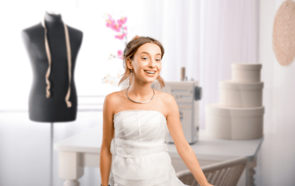 A young woman in a wedding dress standing in front of a sewing machine & mannequin.